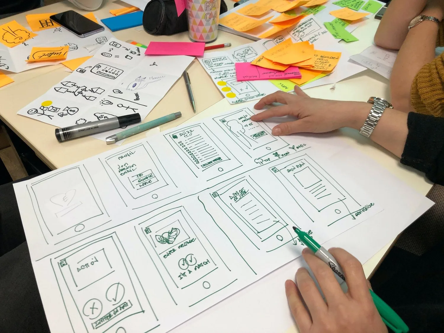 Hand-drawn mobile app wireframes on paper, surrounded by sticky notes and sketches during a design workshop
