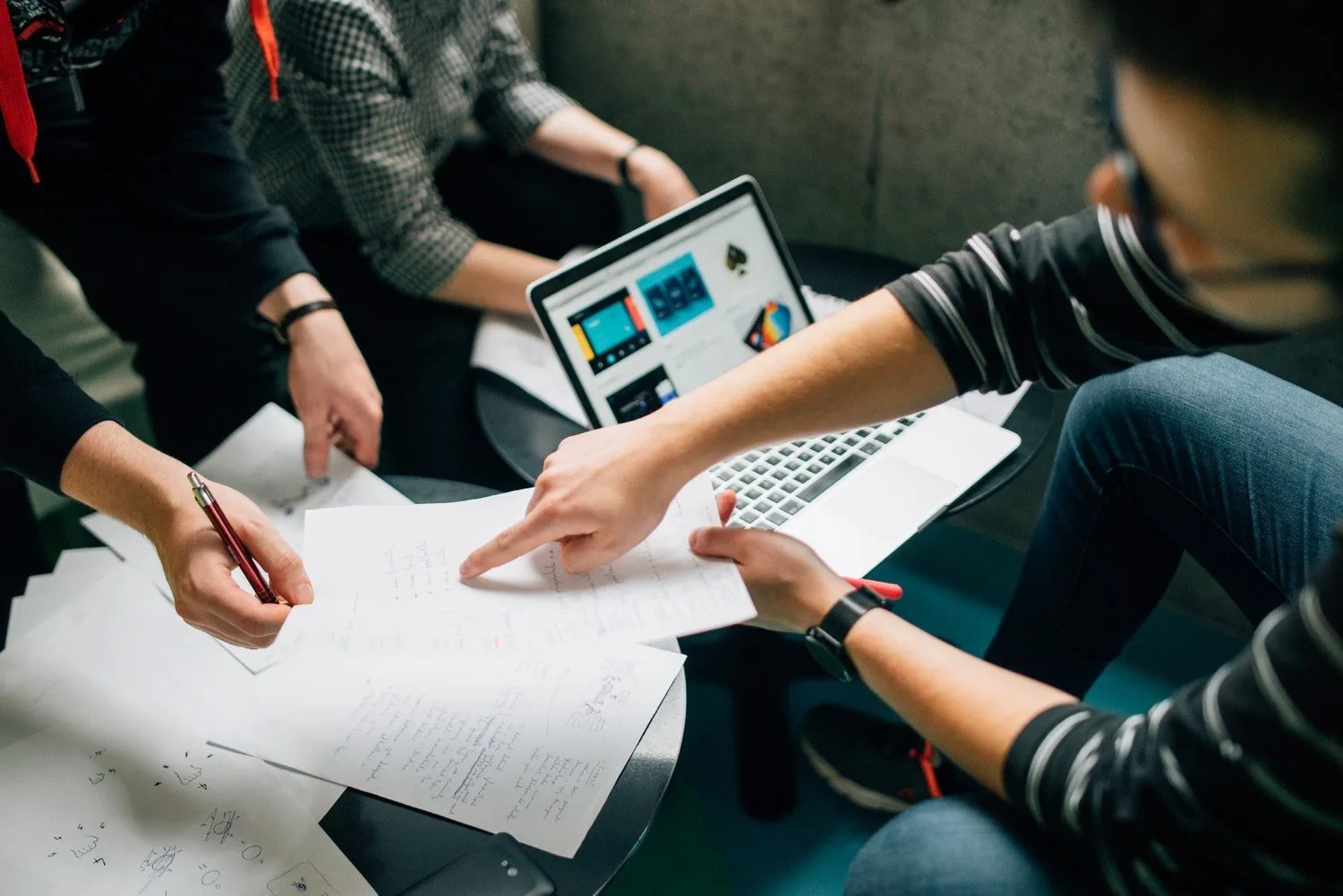 Team members point at handwritten notes on a small table next to a laptop showing app designs