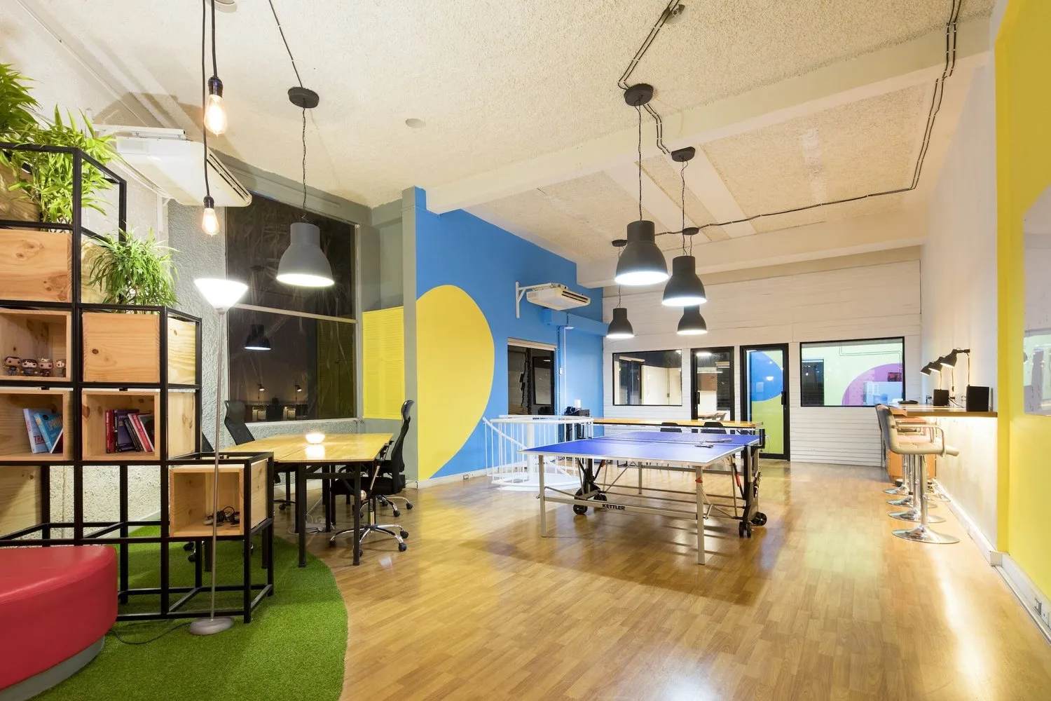 Colourful open-plan startup office with wooden desks, a table tennis table, pendant lamps and shelves with plants