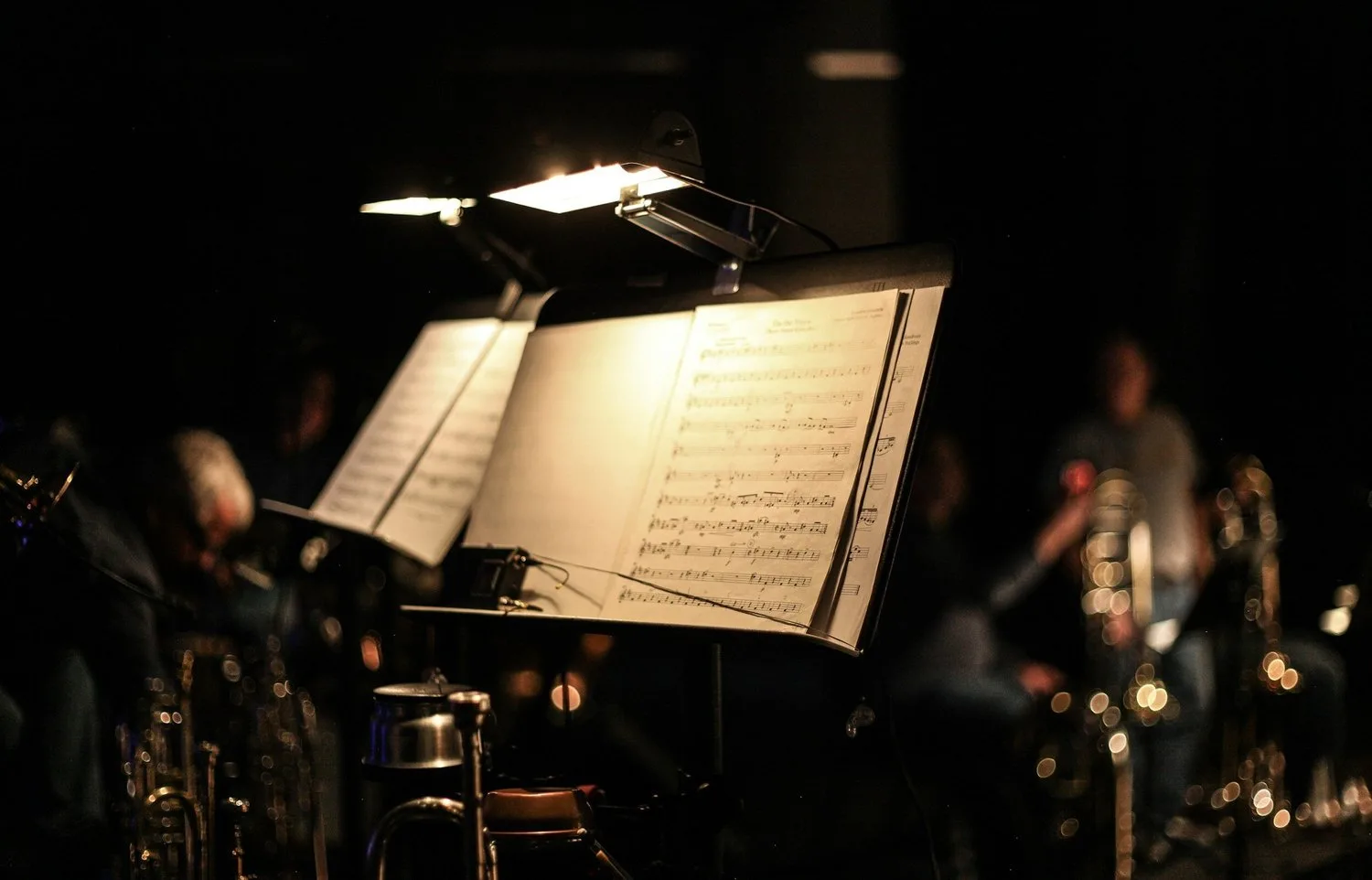 Lit music stands with sheet music in front of blurred musicians and brass instruments on a dark stage