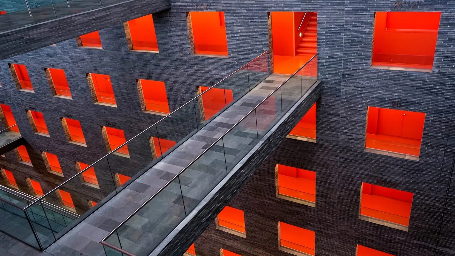 Atrium of a dark brick building with orange-lit window openings and a glass-railed footbridge crossing it