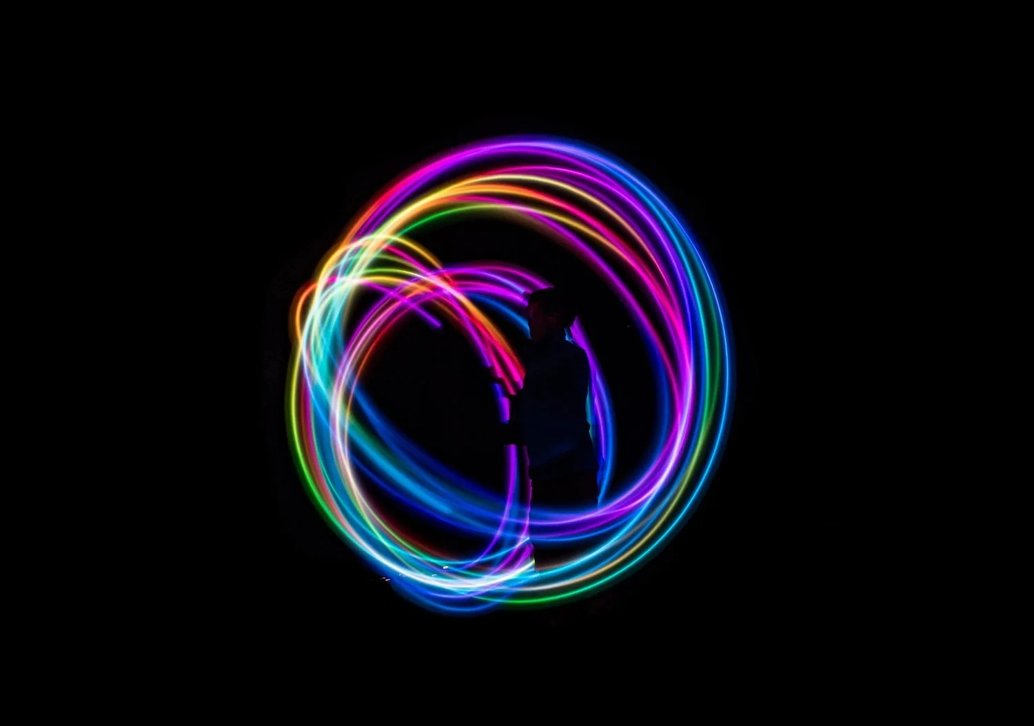 Long-exposure photo of a silhouetted person surrounded by rainbow-coloured light trails in the dark